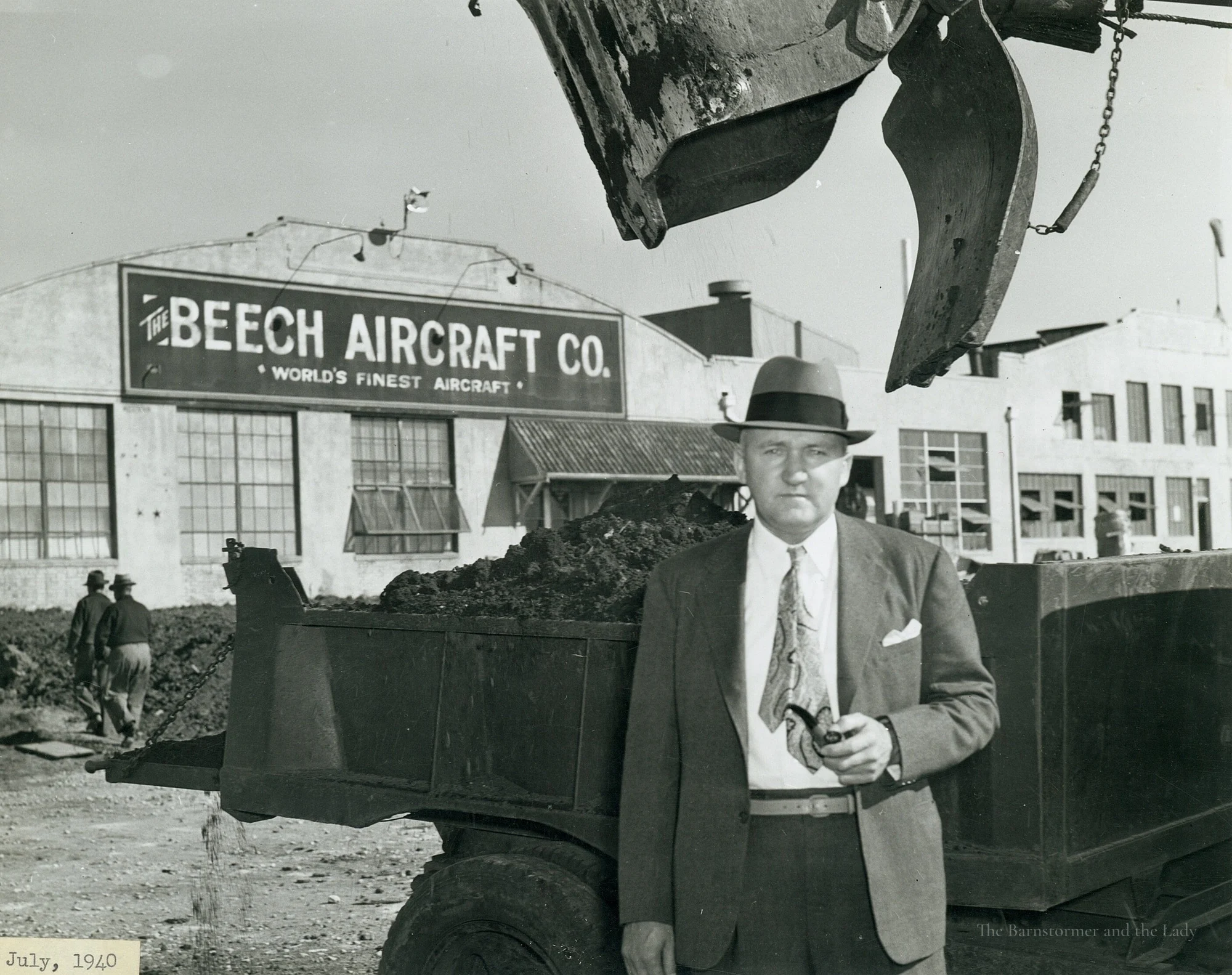 Walter Beech in a hat, holding a pipe, standing in front of a truck outside a building signed The Beech Aircraft Co.