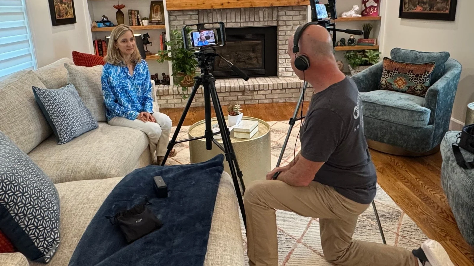 A woman on a sofa faces a camera on a tripod; the camera operator kneels with his back to us
