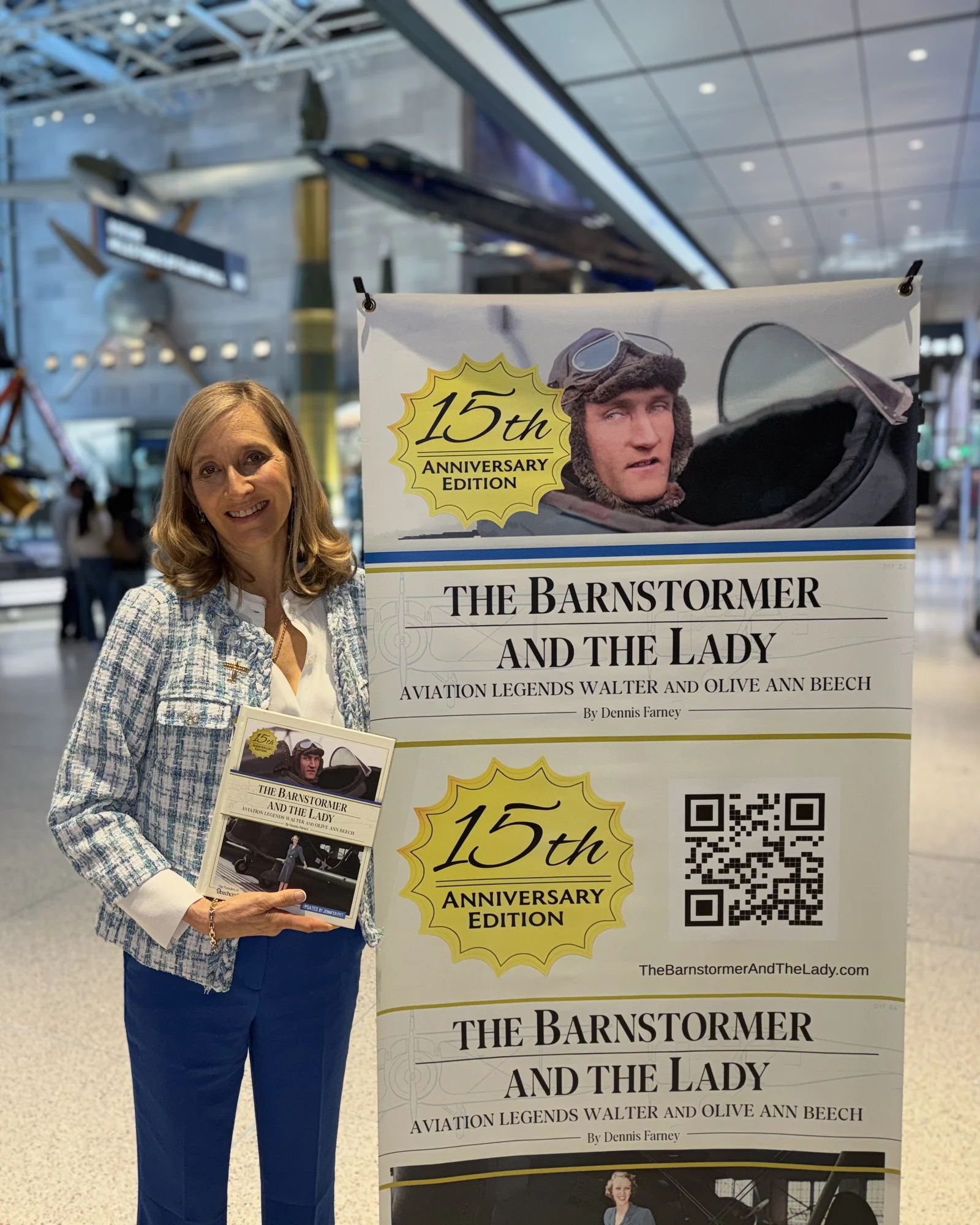 Jennifer Pitt holding the book beside a banner for The Barnstormer and the Lady in a museum hall