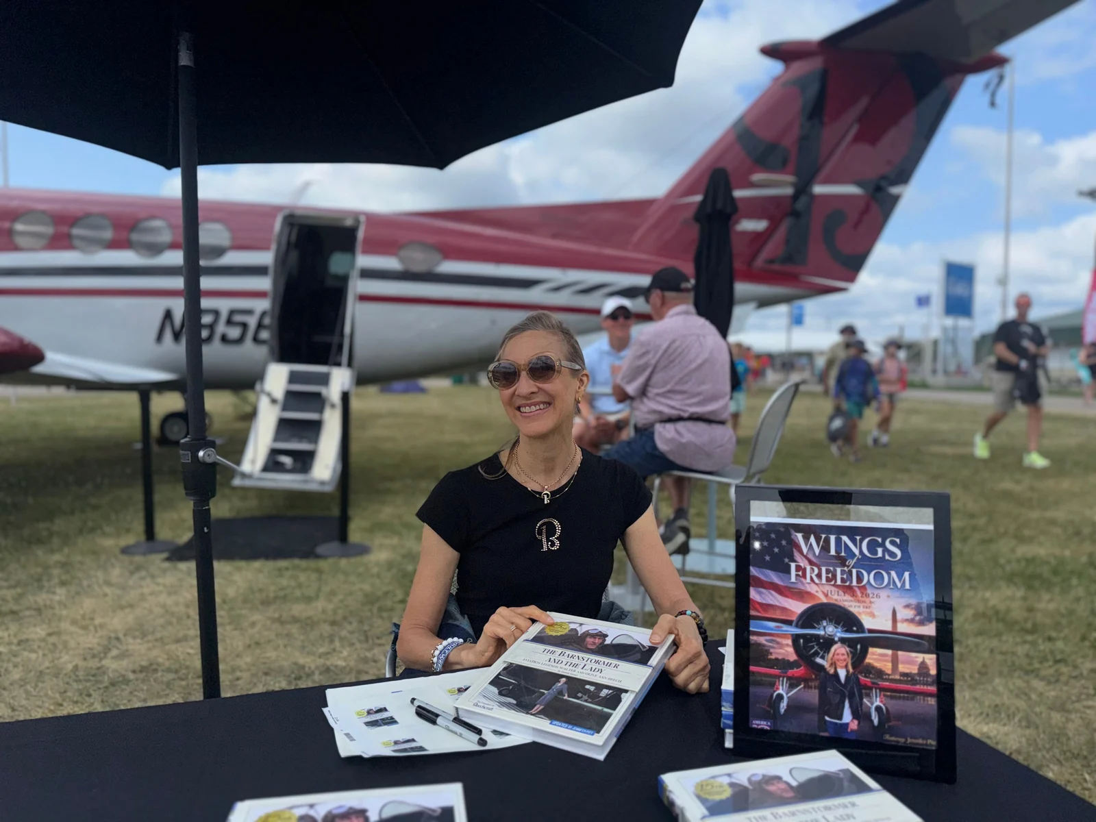 Jennifer Pitt seated at a table under an umbrella, holding the book, with a King Air behind her