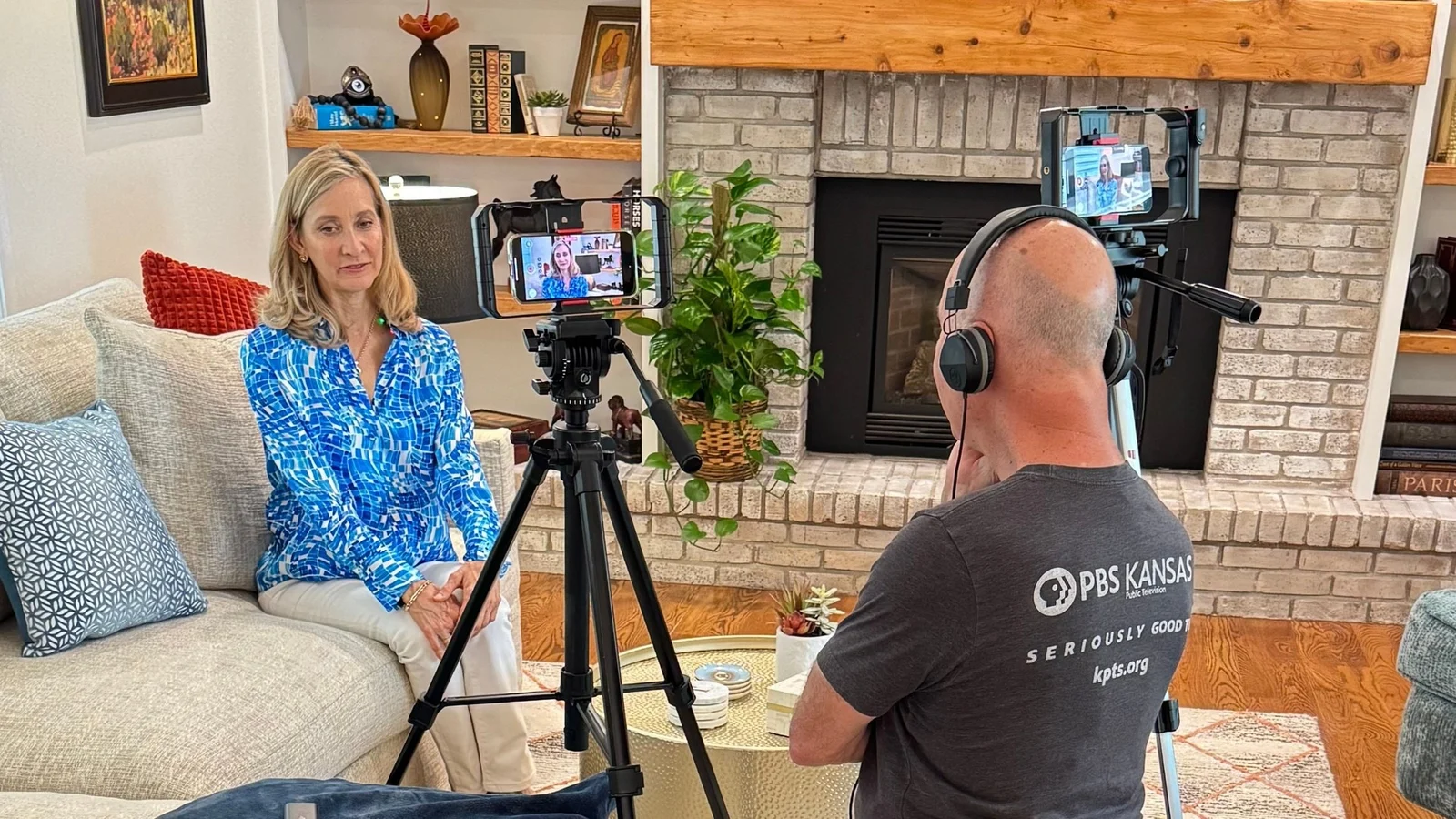 A woman on a sofa is filmed by a camera operator whose shirt reads PBS Kansas