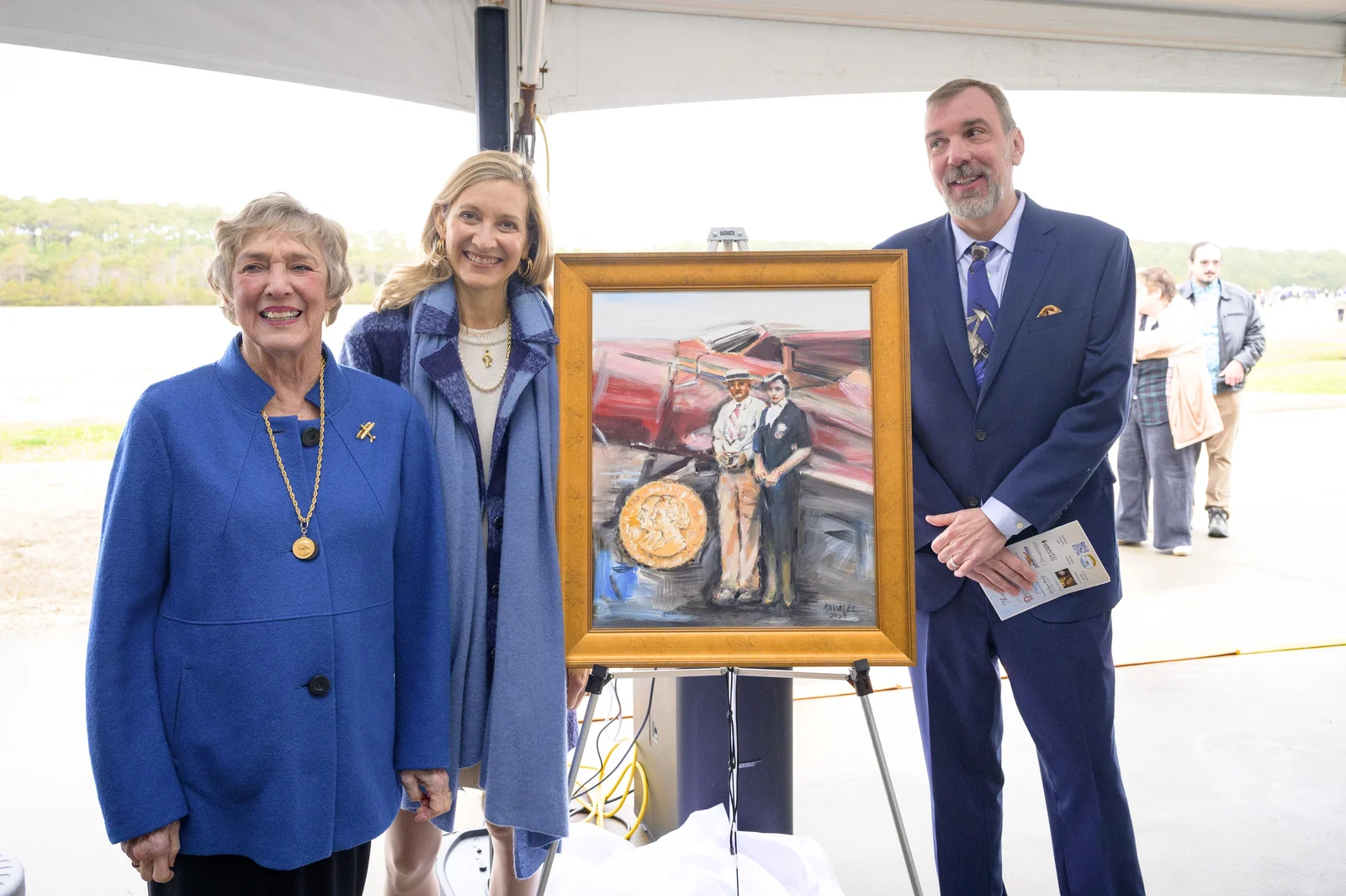 Three people standing beside a framed painting on an easel under an outdoor tent