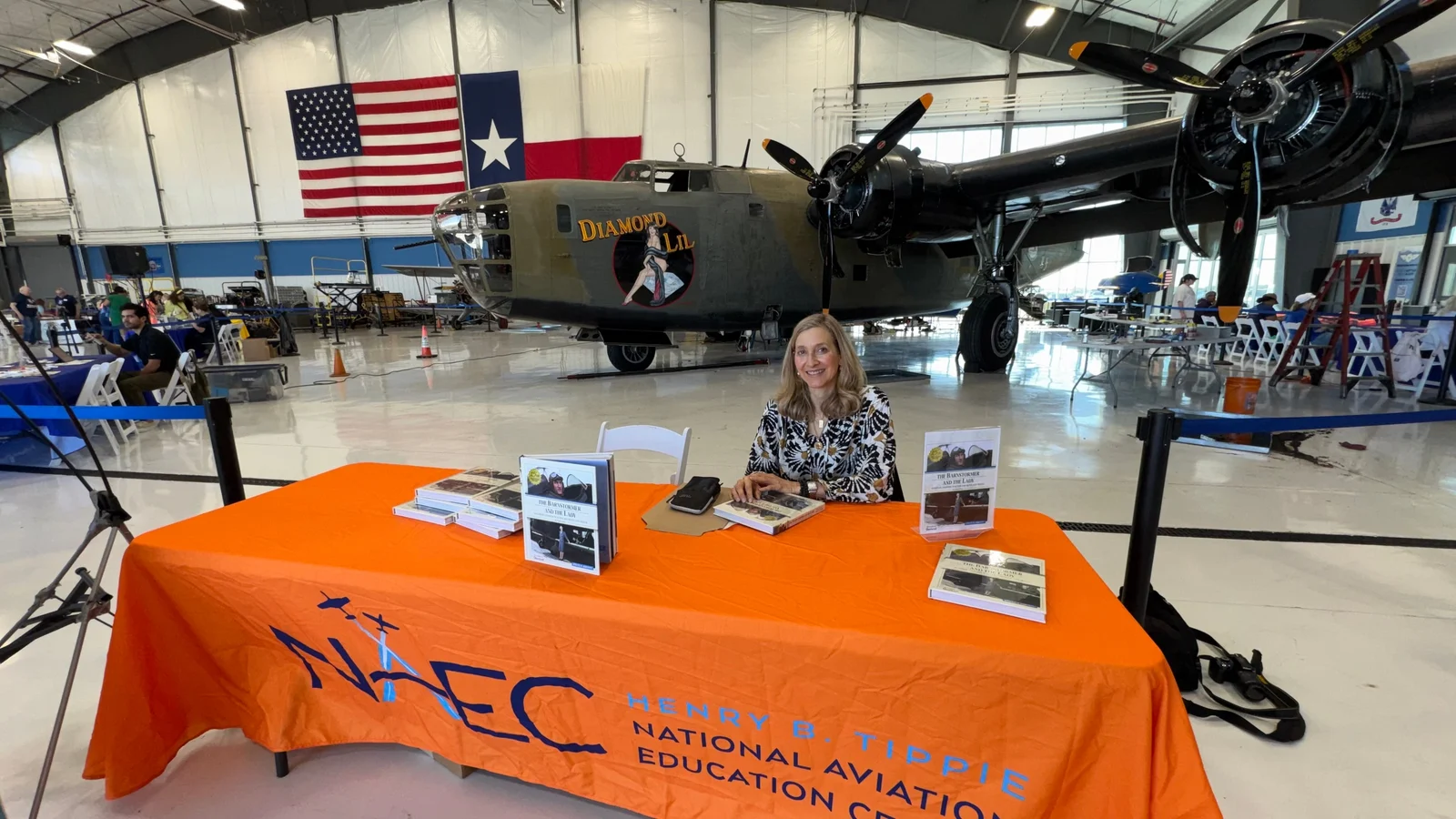 Jennifer Pitt seated at a table of books in an aircraft hangar, in front of a vintage bomber and the United States and Texas flags